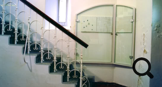 Treppenhaus mit weißem Geländer und schwarzem Handlauf, daneben ein Glaskasten an der Wand.
